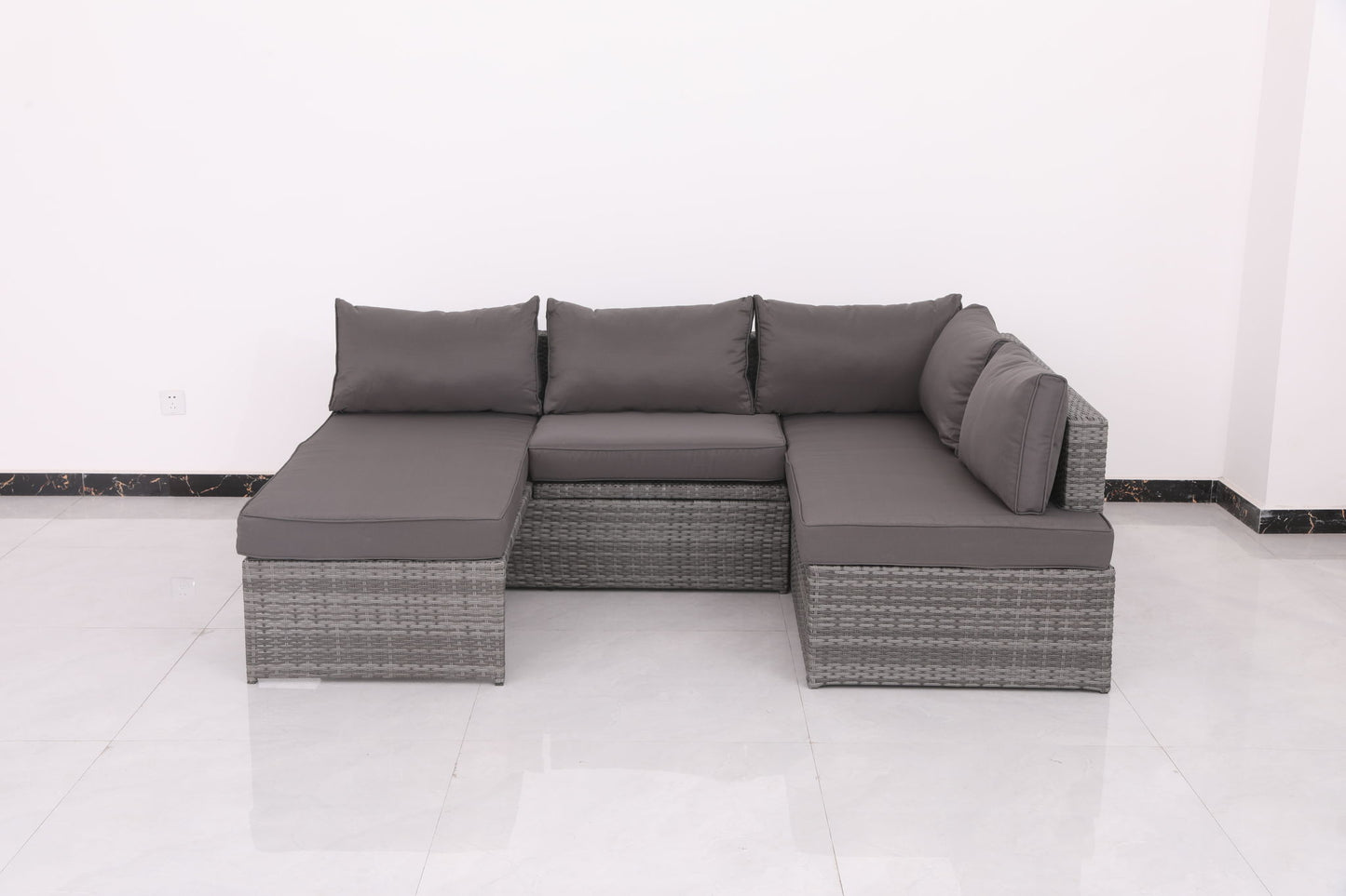 The Patio Sofa With Cushions - Gray is an L-shaped wicker sectional with dark gray cushions, three backrest cushions, and a right-side armrest. It's shown against a white wall on a glossy white tile floor.