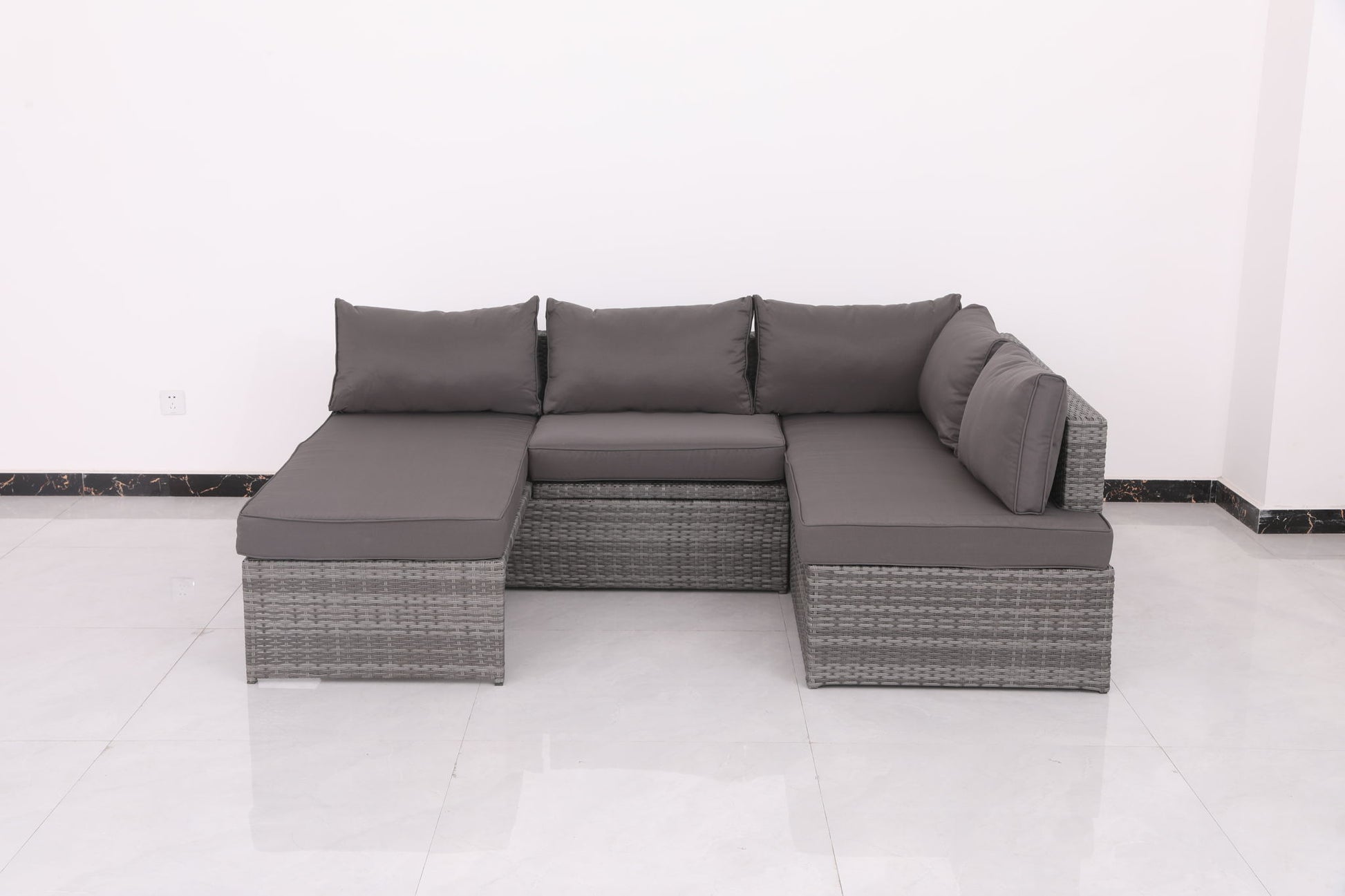The Patio Sofa With Cushions - Gray is an L-shaped wicker sectional with dark gray cushions, three backrest cushions, and a right-side armrest. It's shown against a white wall on a glossy white tile floor.