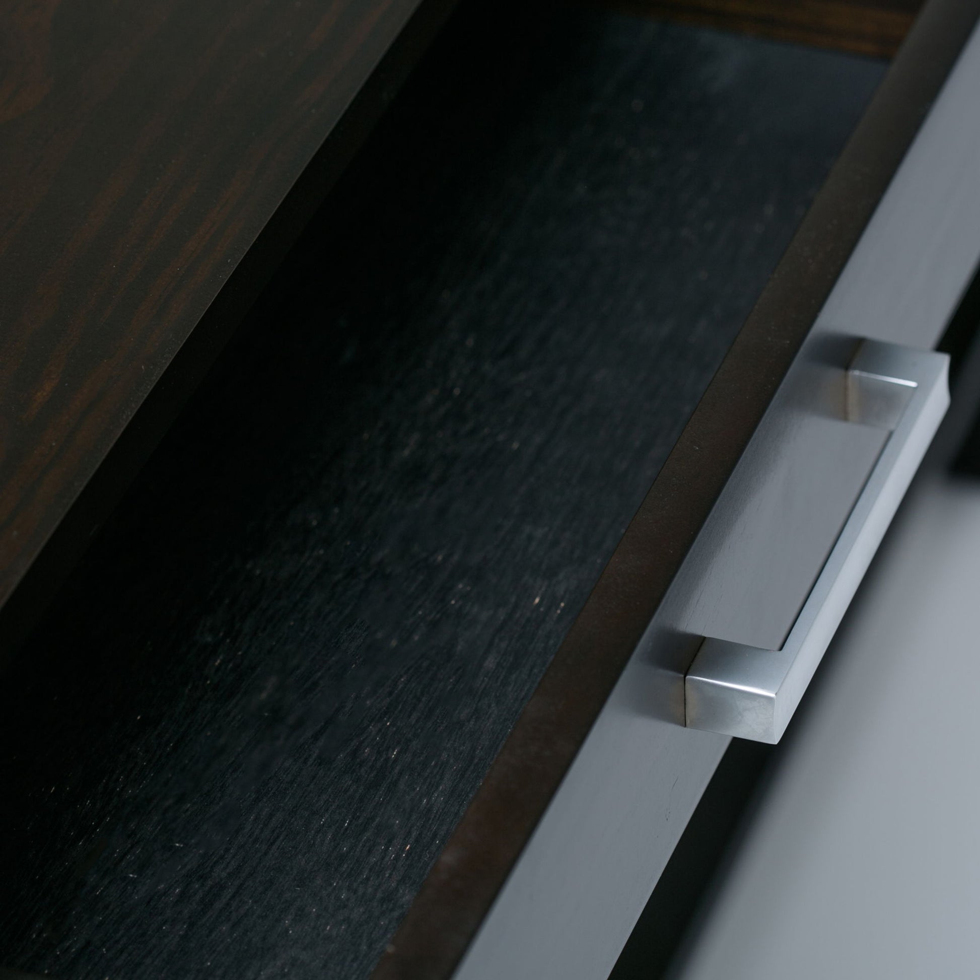 A close-up view of a partially open drawer with a silver rectangular handle on the Kitchener - Handcrafted Table, shown from above. The dark wooden drawer appears empty.