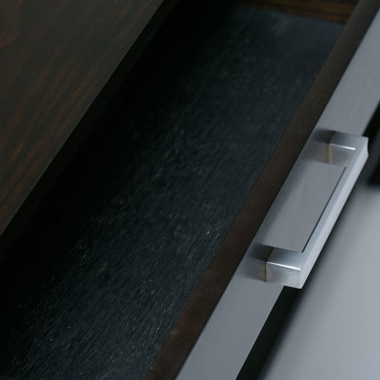 A close-up view of a partially open drawer with a silver rectangular handle on the Kitchener - Handcrafted Table, shown from above. The dark wooden drawer appears empty.