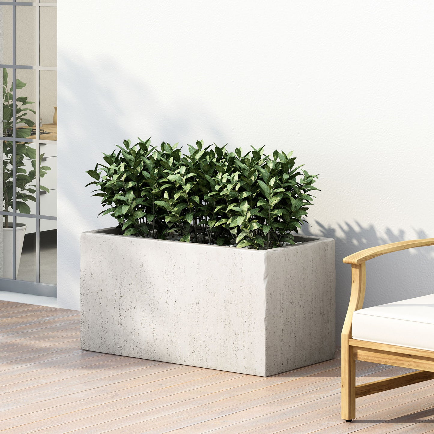 The Modern Outdoor Planter with dense green foliage sits on a wooden patio floor beside a wooden outdoor chair with white cushions, set against a white wall.