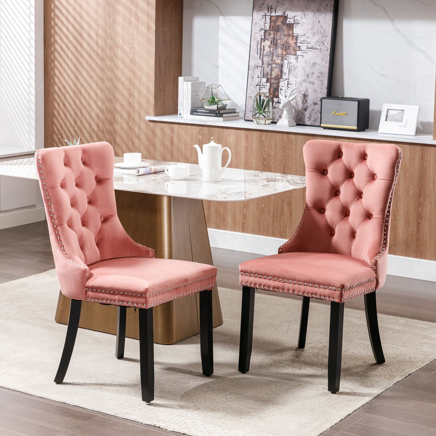 Two Nikki modern tufted velvet dining chairs with wood legs and nailhead trim sit before a marble table in a bright, wood-paneled room with shelving and decor accents.