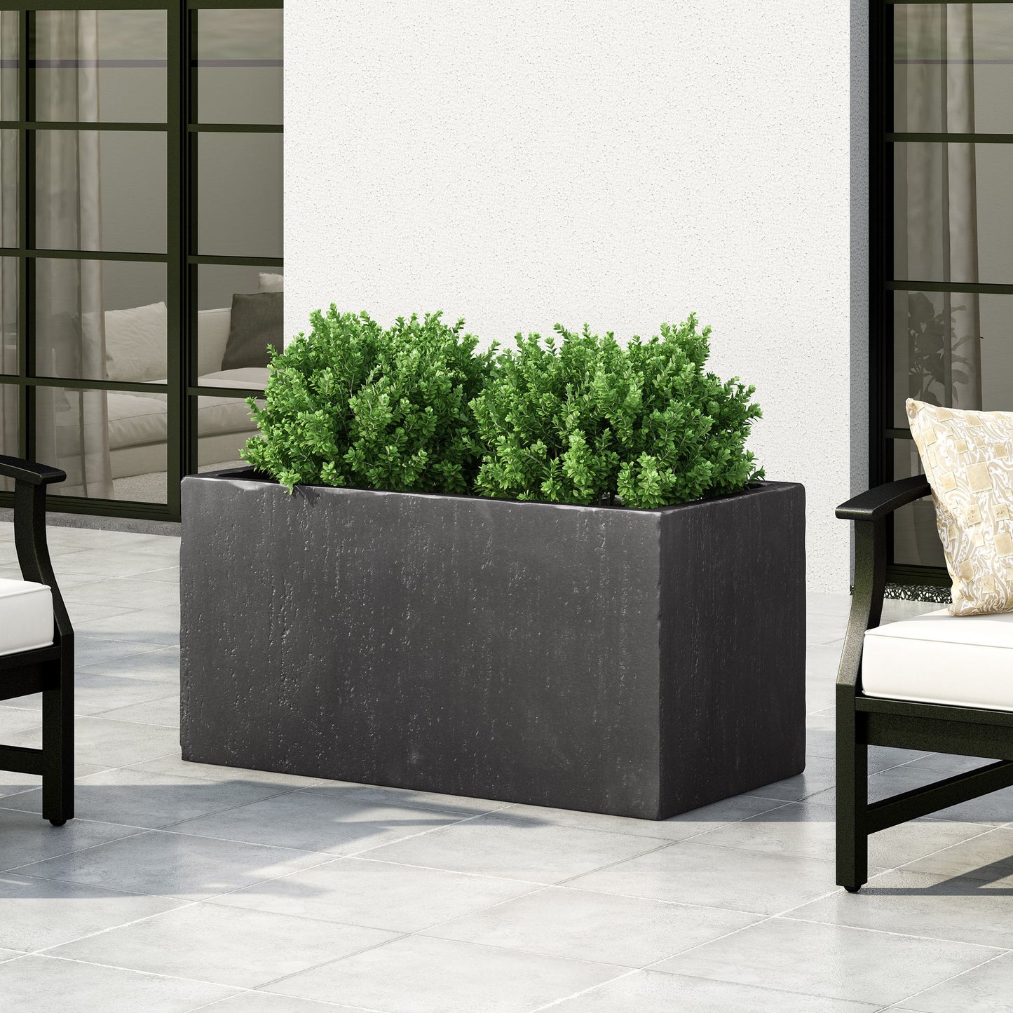 A modern outdoor patio features the Modern Outdoor Planter in black, filled with green shrubs and set between two cushioned chairs on a tiled floor in front of tall windows.