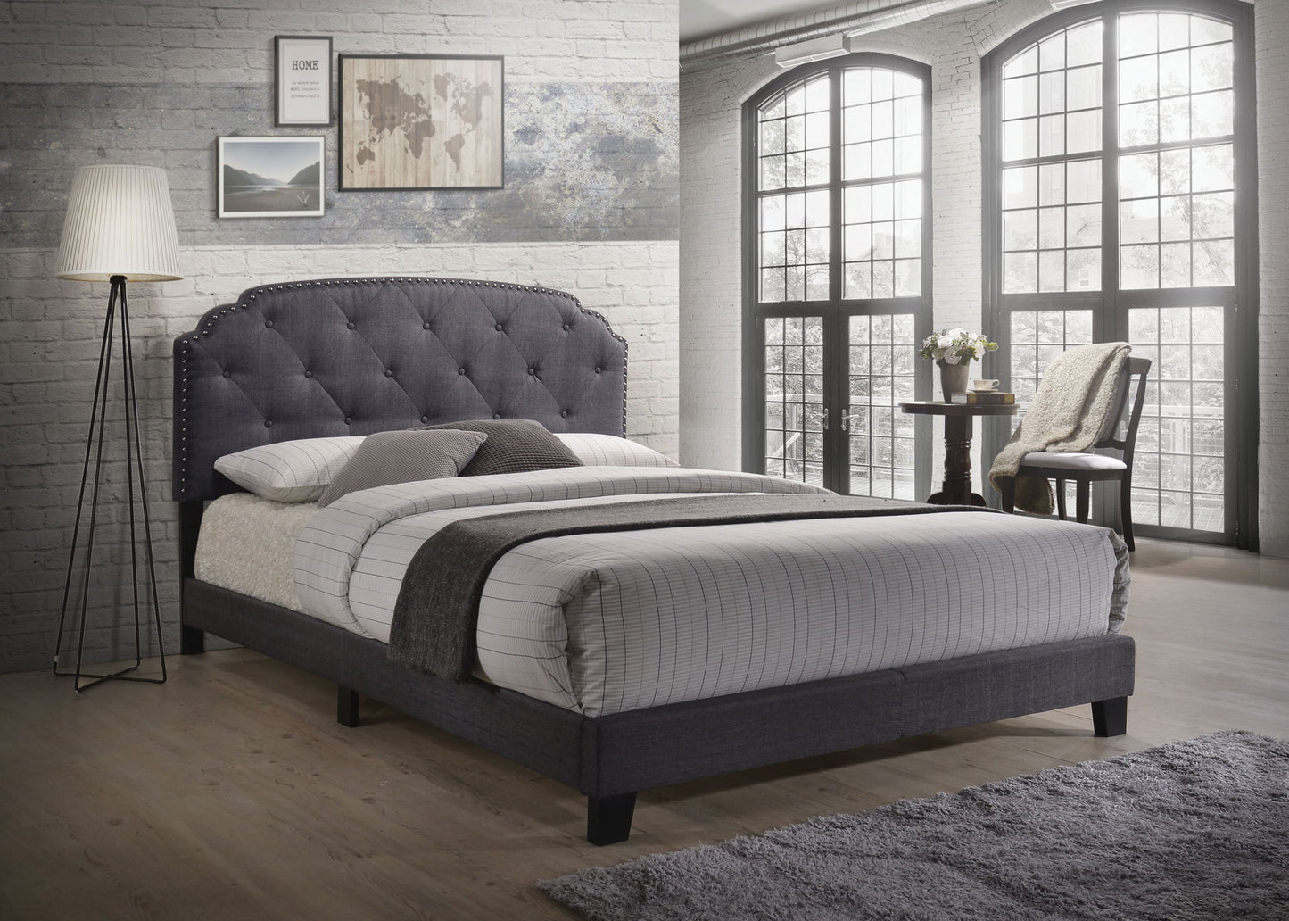 A modern bedroom featuring the Tradilla - Queen Bed in gray, white and gray bedding, a floor lamp, wall art, large arched windows, light wood flooring, brick walls, a small round table with flowers, and a chair draped with a blanket.