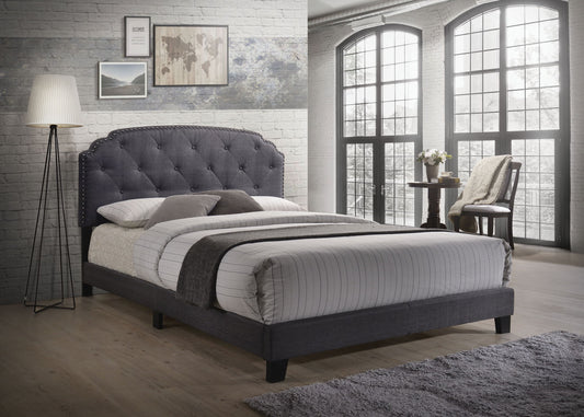 A modern bedroom featuring the Tradilla - Queen Bed in gray, white and gray bedding, a floor lamp, wall art, large arched windows, light wood flooring, brick walls, a small round table with flowers, and a chair draped with a blanket.