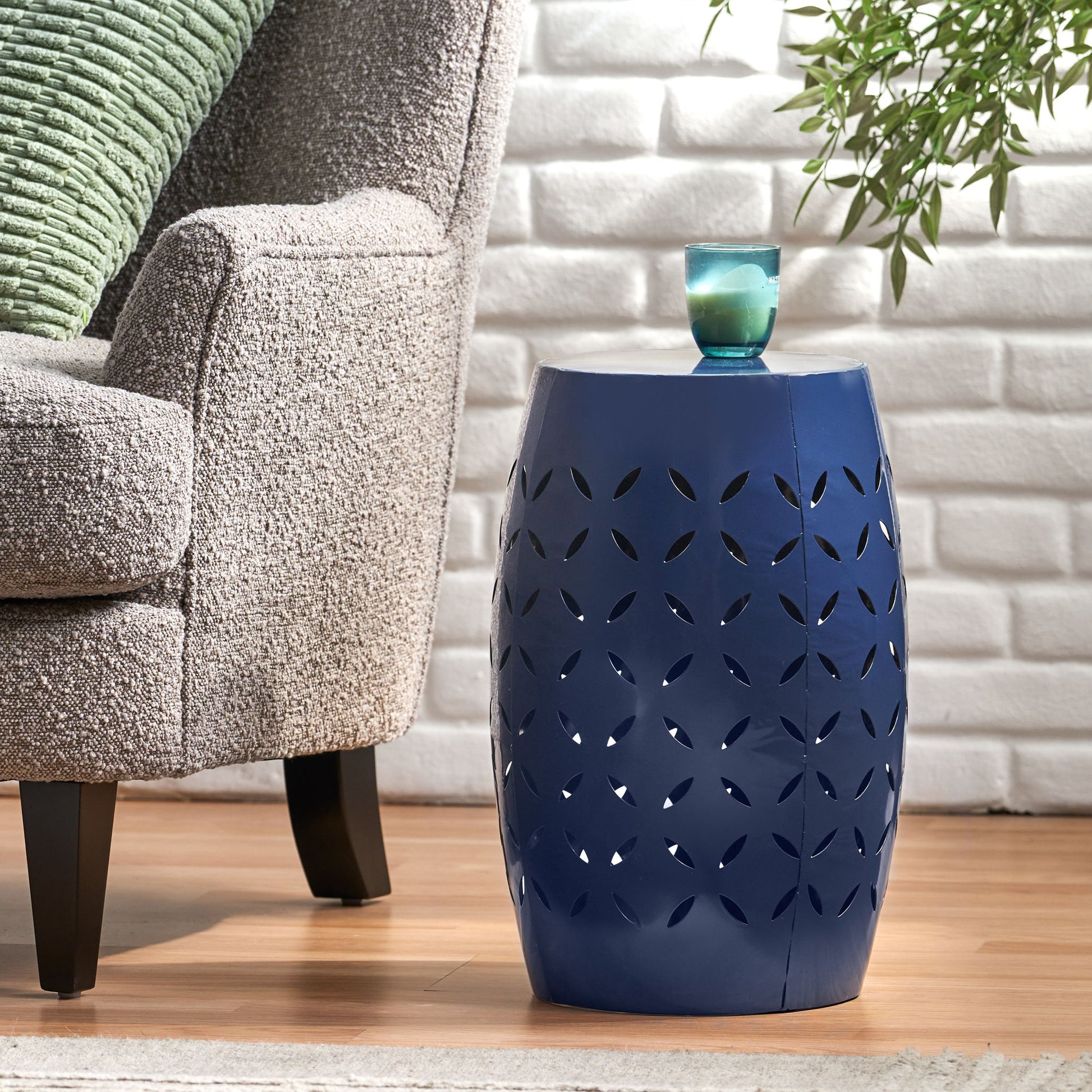 The 12" Outdoor Lace Cut Side Table in blue holds a small glass cup. It stands on a wooden floor beside a gray upholstered chair, in front of a white brick wall with green plant leaves peeking into view.