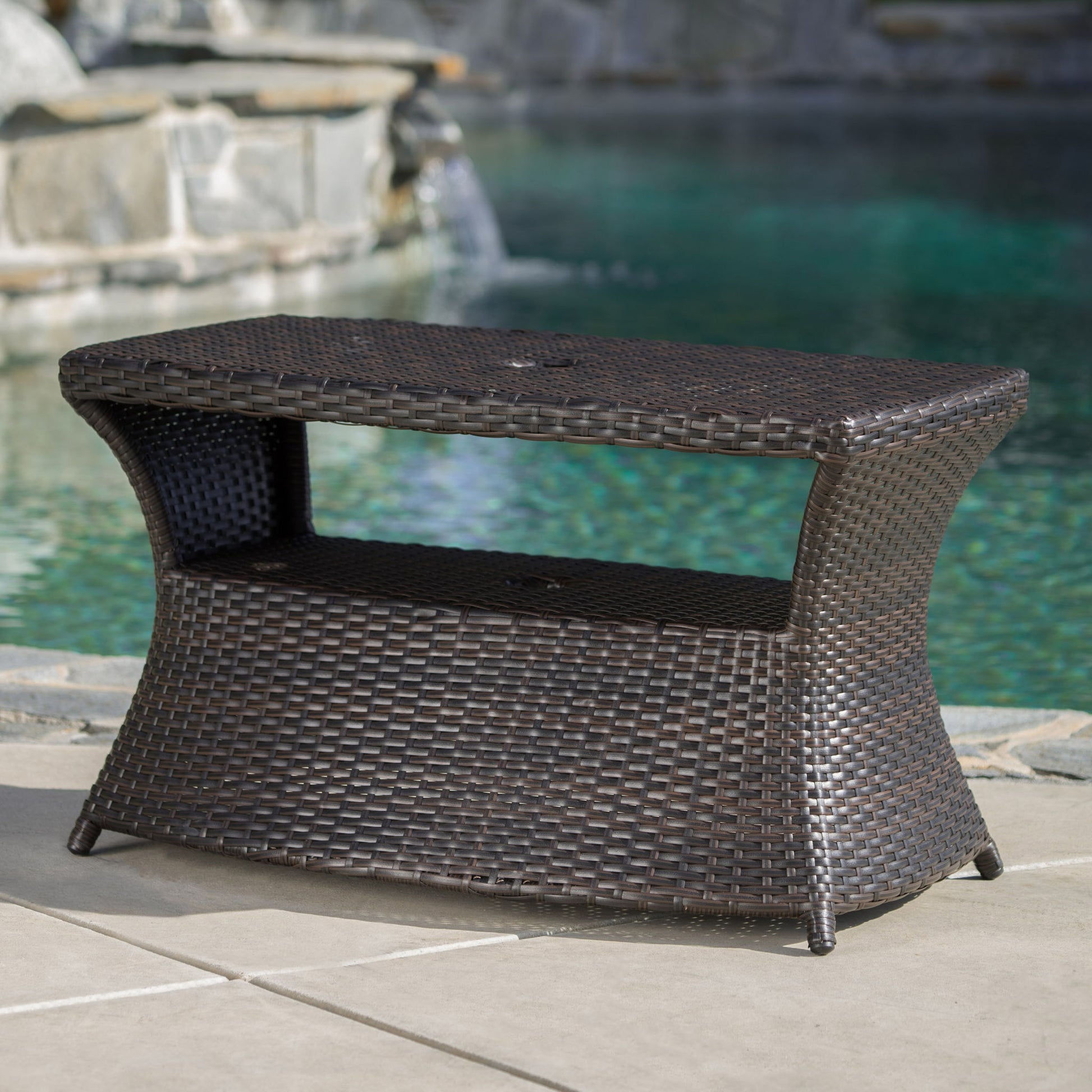 The Berkeley Chaise Lounge Side Table, in dark brown wicker with a shelf, is shown on a patio beside a pool and stone water feature.
