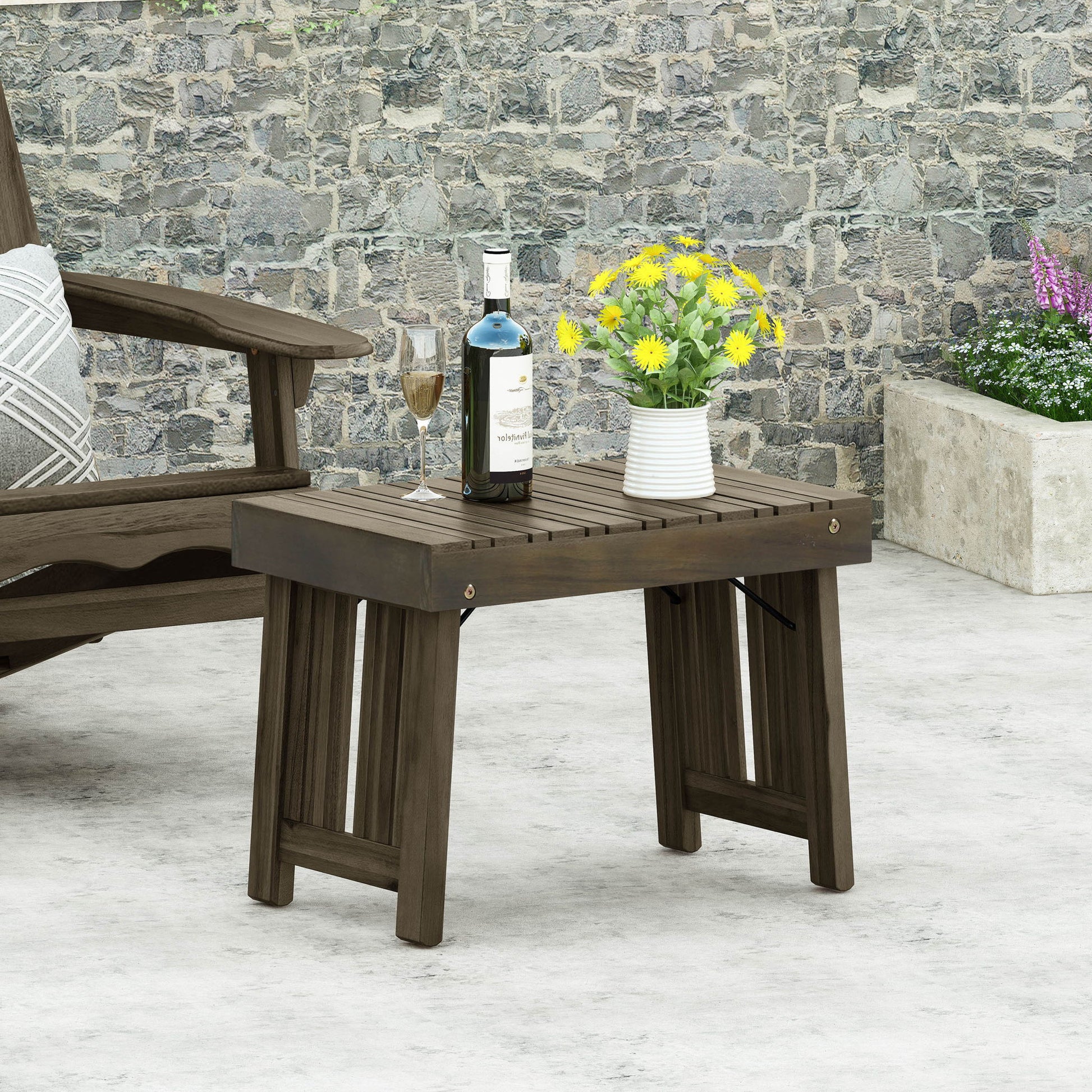 The Temptation Outdoor Foldable Side Table With Slat Design holds a wine bottle, wine glass, and a vase of yellow flowers. A cushioned chair is partly visible on the left with a stone wall in the background.