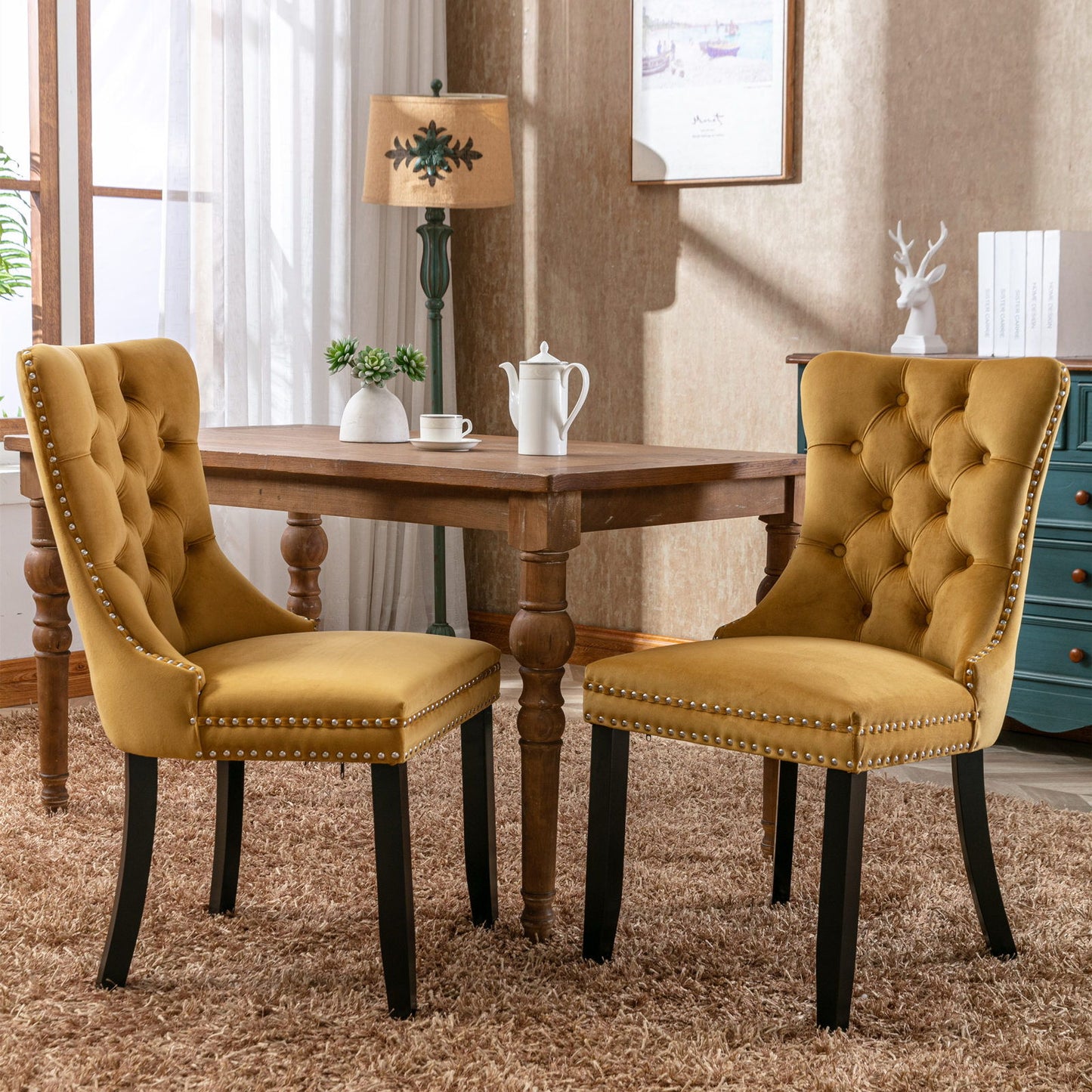 Two Nikki Modern Velvet Tufted Dining Chairs with nailhead trim and wood legs are placed around a wooden table with a white teapot and cup, in a sunlit room featuring beige walls, a lamp, and framed artwork.
