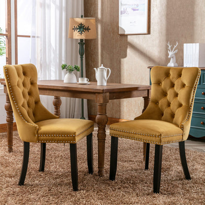 Two Nikki Modern Velvet Tufted Dining Chairs with nailhead trim and wood legs are placed around a wooden table with a white teapot and cup, in a sunlit room featuring beige walls, a lamp, and framed artwork.