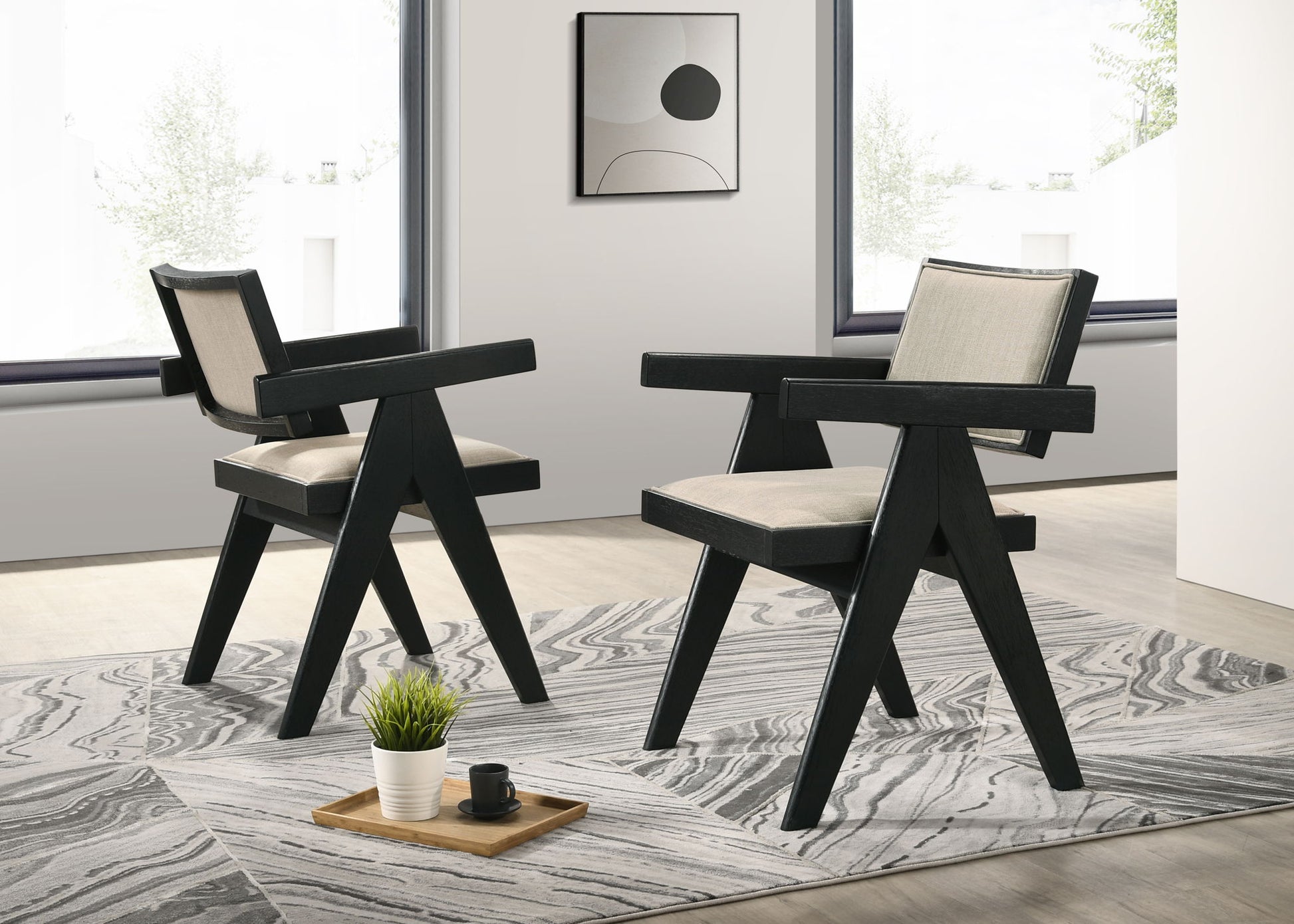 The Jasper 3 Piece Arm Chairs and Console Table (Ebony Black) set features modern angular chairs on a patterned rug in a bright, minimalist room with large windows, abstract wall art, and a tray with plant and cup.