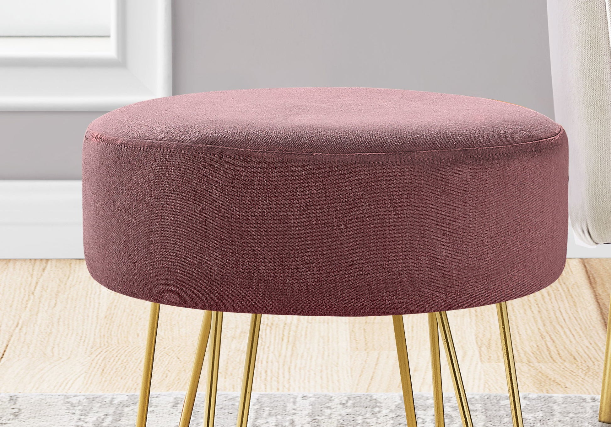 A contemporary round pouf with pink velvet upholstery and gold metal legs sits on a wooden floor beside a light rug and gray wall.