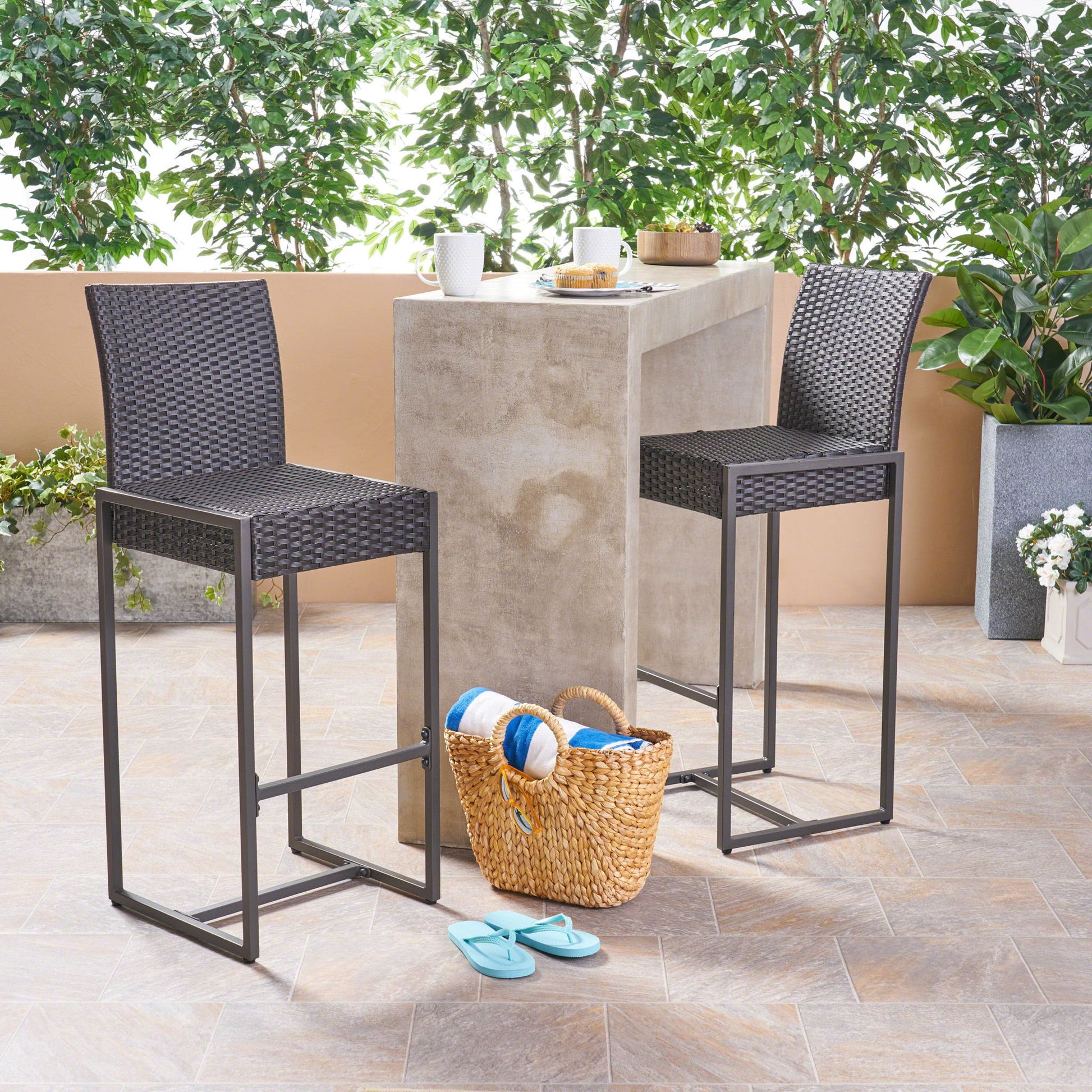 A set of Outdoor Barstool With Hand Crafted Details and a concrete bar table stand on a tiled patio, with a basket holding towels and flip-flops nearby. Potted plants and trees create a lush, green backdrop.