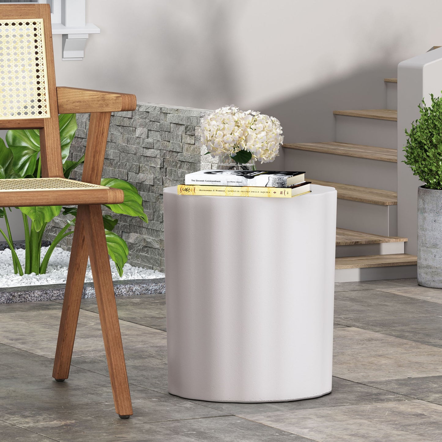 A modern interior features a light wood chair, the Outdoor End Table With Soft Wave Edge in Antique White holding books and white flowers, stone flooring, stairs, and green plants by a stone wall.