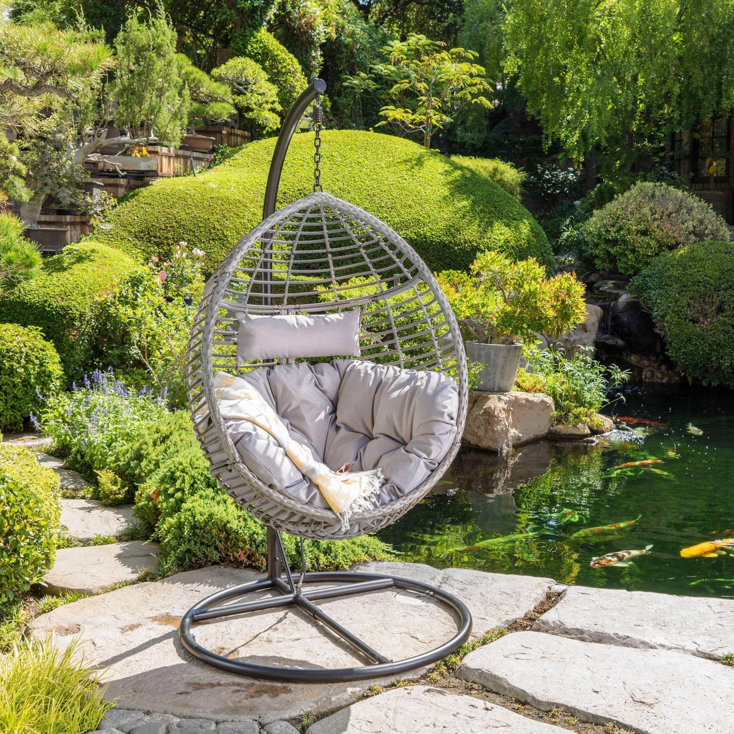 A Hanging Basket Chair With Metal Frame And Seat Cushion sits on a stone patio, overlooking a lush garden and koi pond. Surrounded by green bushes, flowers, and trees, the tranquil outdoor space invites relaxation.