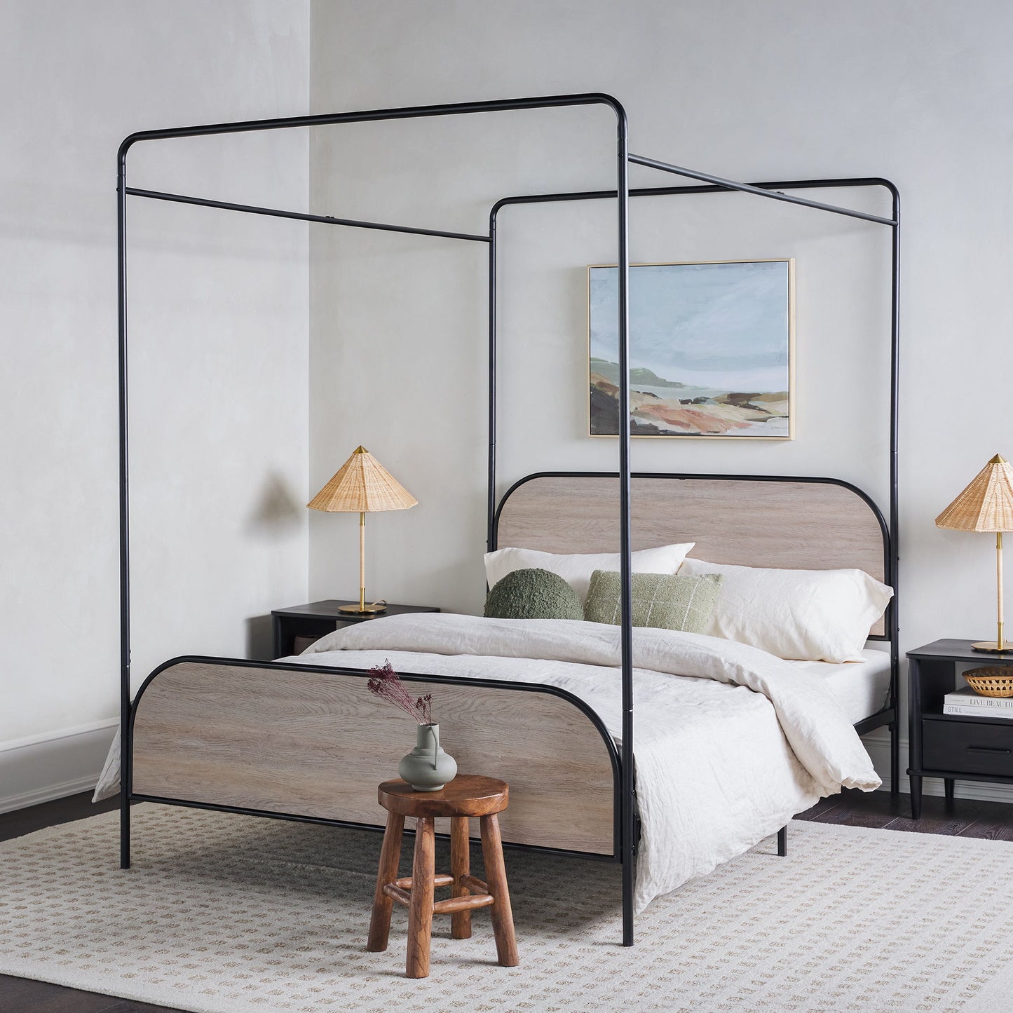A modern bedroom featuring the Modern Canopy Bed Frame with a black metal design, light wood headboard and footboard, white bedding, green pillows, bedside tables with matching lamps, a landscape painting, and a wooden stool with a vase.