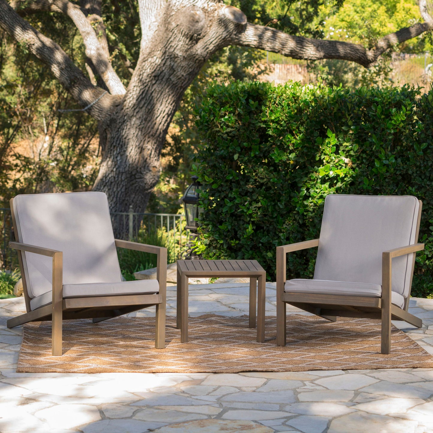 The Celestaria 3 Piece Outdoor Chat Set features cushioned Adirondack chairs with a side table, arranged on a patio rug with lush greenery in the background.