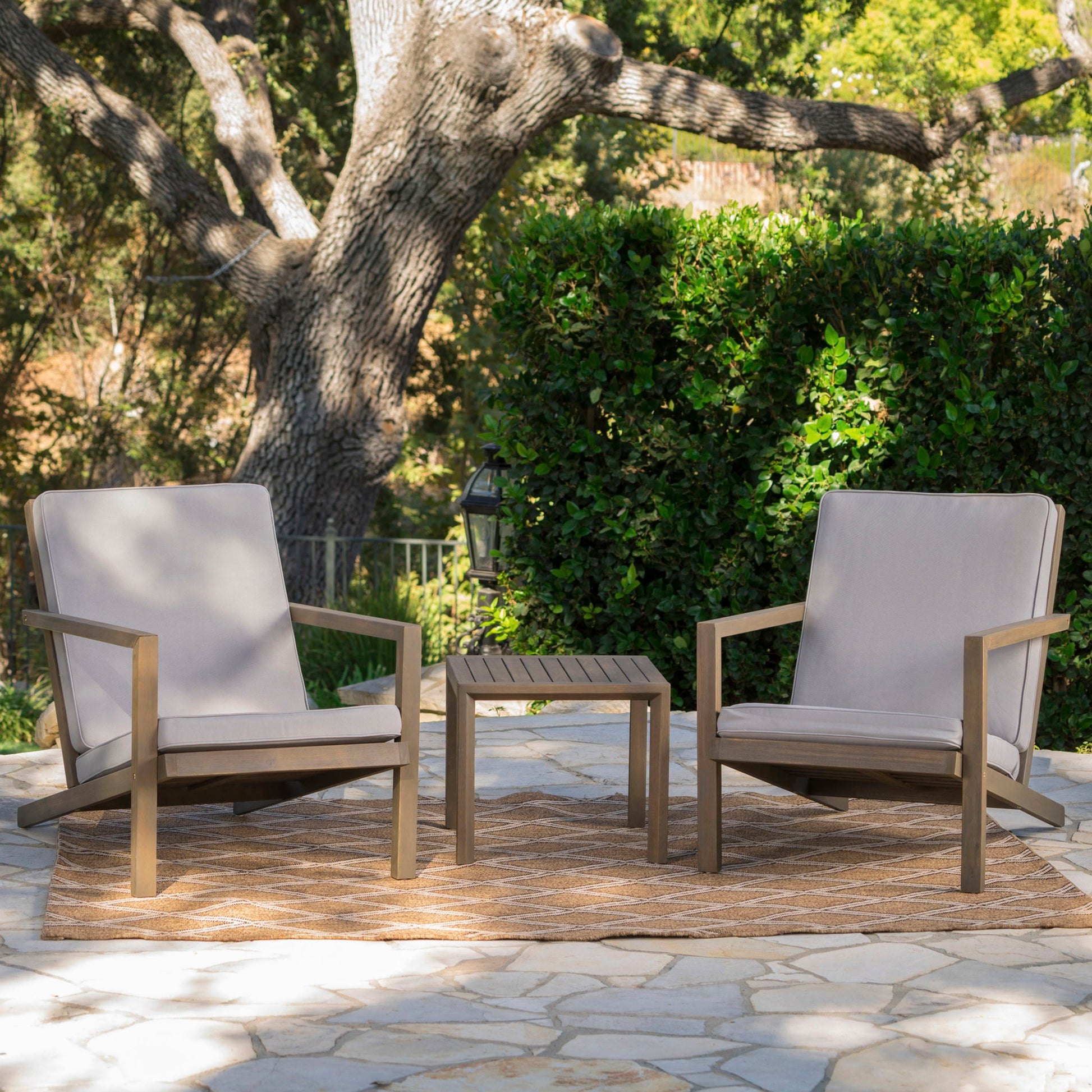 The Celestaria 3 Piece Outdoor Chat Set features cushioned Adirondack chairs with a side table, arranged on a patio rug with lush greenery in the background.
