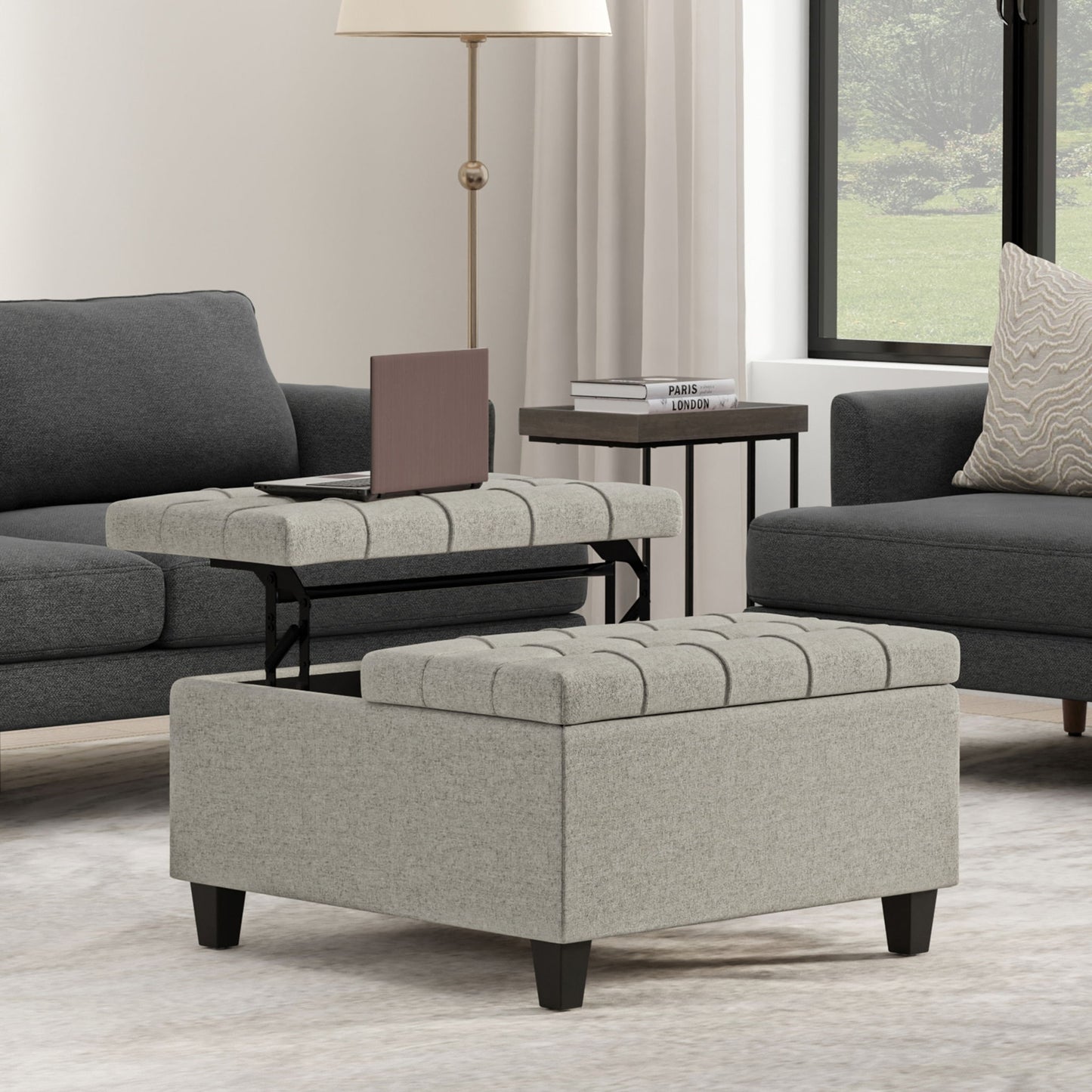 A modern living room showcases the Harrison Upholstered Coffee Table Storage Ottoman with a lift-top tray, set between a dark gray sofa and a side table with books and a lamp; greenery is visible through the window.