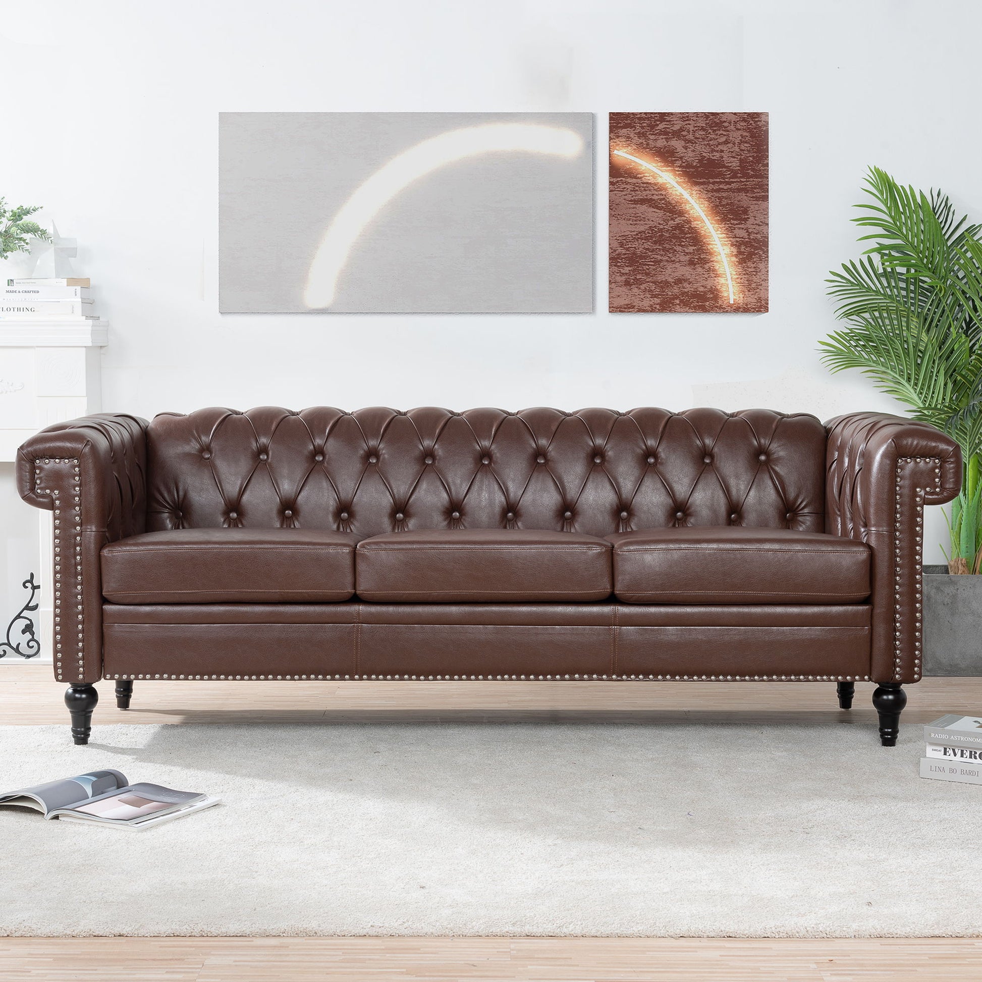 A Traditional Square Arm Removable Cushion 3 Seater Sofa with brown tufted leather and nailhead trim sits on a light rug, beneath two abstract art pieces. A green potted plant is on the right, with an open magazine on the floor in a modern living room.