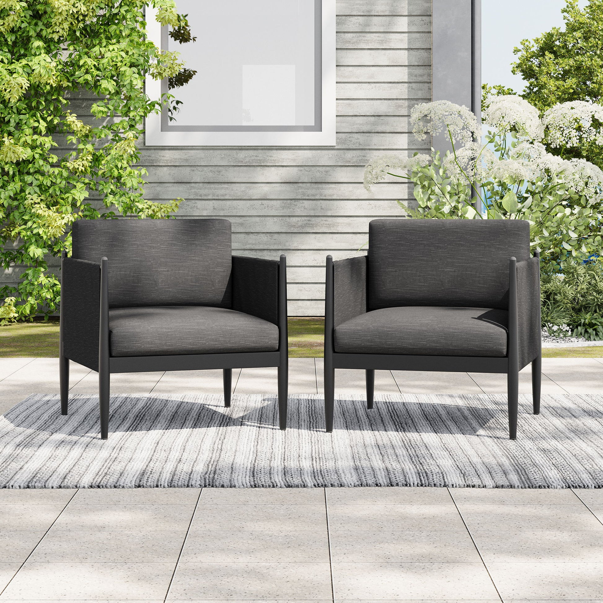 Two Dawson Club Chairs with dark cushions sit side by side on a gray outdoor rug, placed on a tiled patio with green plants and a gray building in the background.