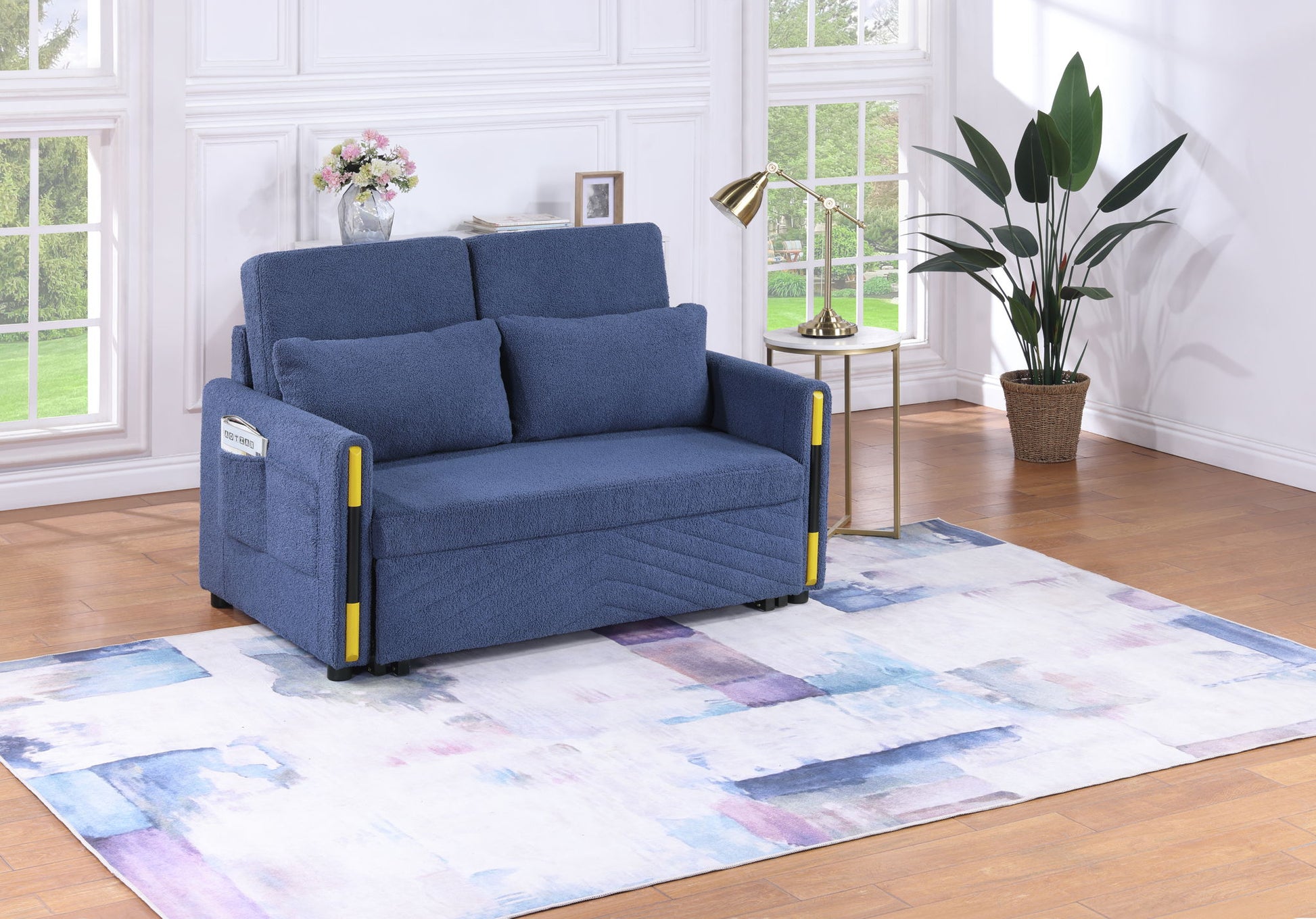 A modern living room featuring the Percy - Teddy Velvet Sleeper Loveseat with dual-pull sleeper design, two cushions, a side pocket, a white and blue abstract rug, a gold floor lamp, a potted plant, large windows, white walls, and wooden floors.