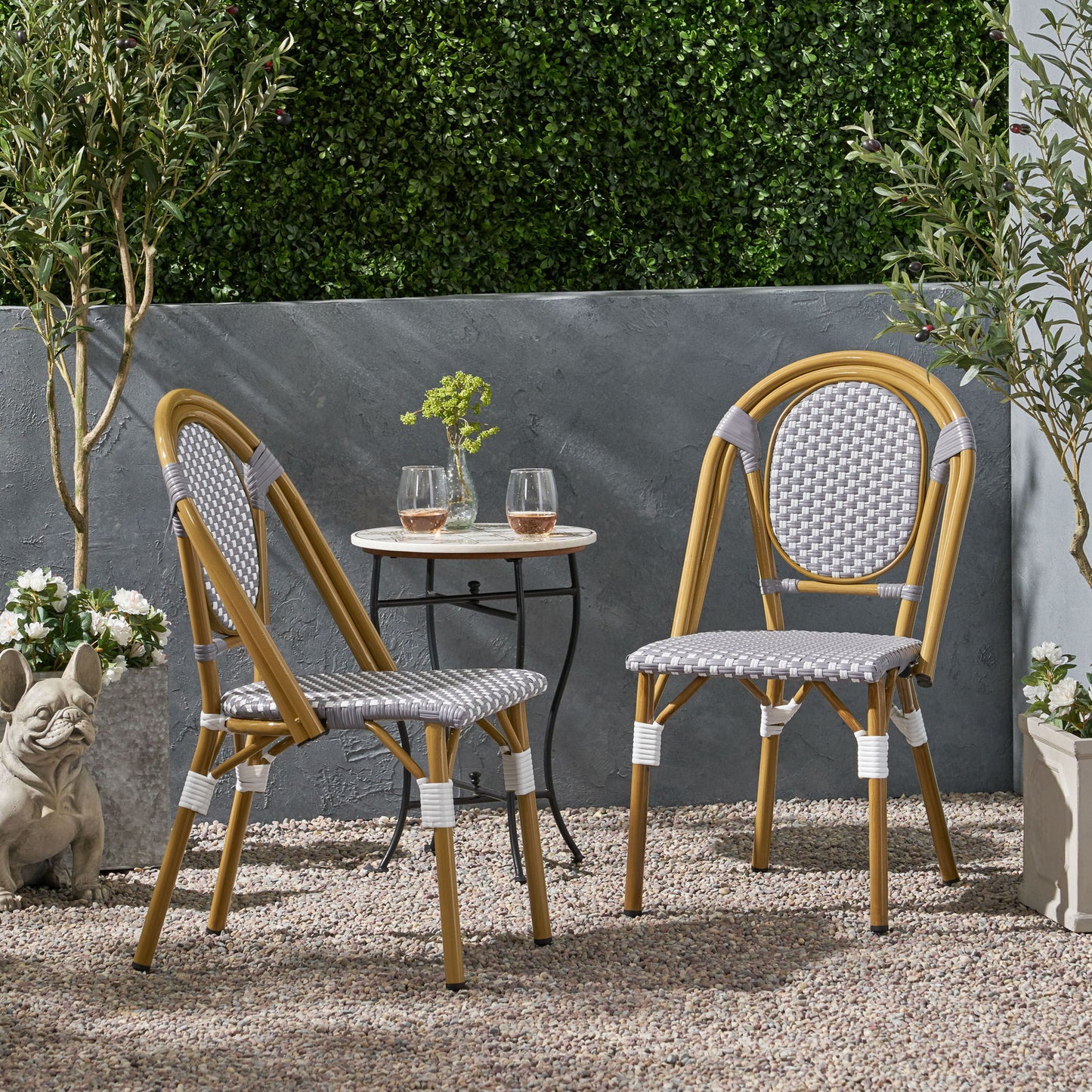 The Outdoor Rattan And Aluminum French Bistro Chair Set with a small round table and two glasses of rosé sits on a gravel patio, surrounded by potted plants, a green hedge, and a decorative stone dog statue.