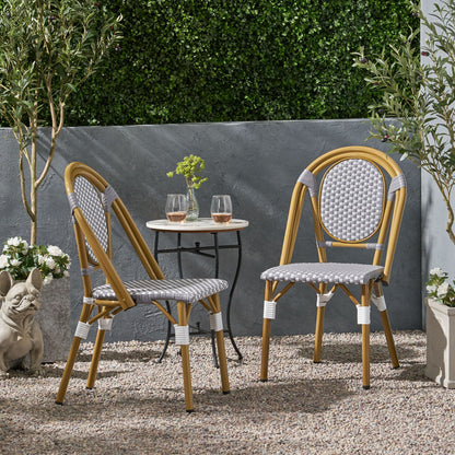The Outdoor Rattan And Aluminum French Bistro Chair Set with a small round table and two glasses of rosé sits on a gravel patio, surrounded by potted plants, a green hedge, and a decorative stone dog statue.