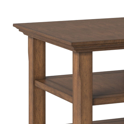 Close-up of the Acadian - Transitional End Table, showcasing its rectangular top, two lower shelves, natural brown finish, and sturdy legs. The table is set against a plain white background.