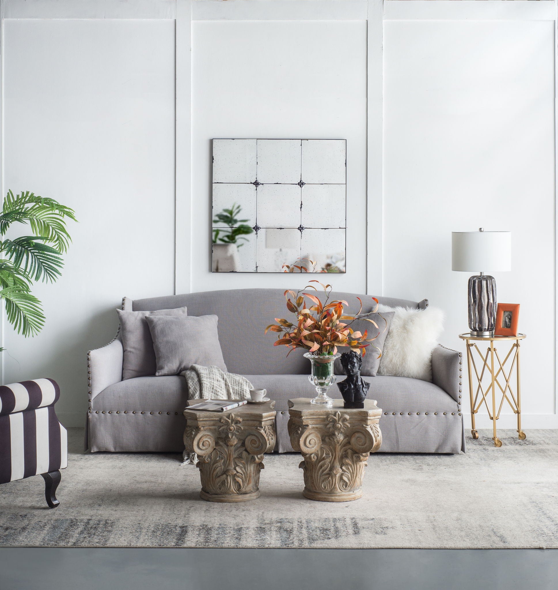 A modern living room with a gray sofa, neutral pillows, stone pedestal coffee tables, a plant, striped chair, gold side table with lamp and photo, and a Traditional Decor Style Antique Glass Panel Mirror above the sofa.