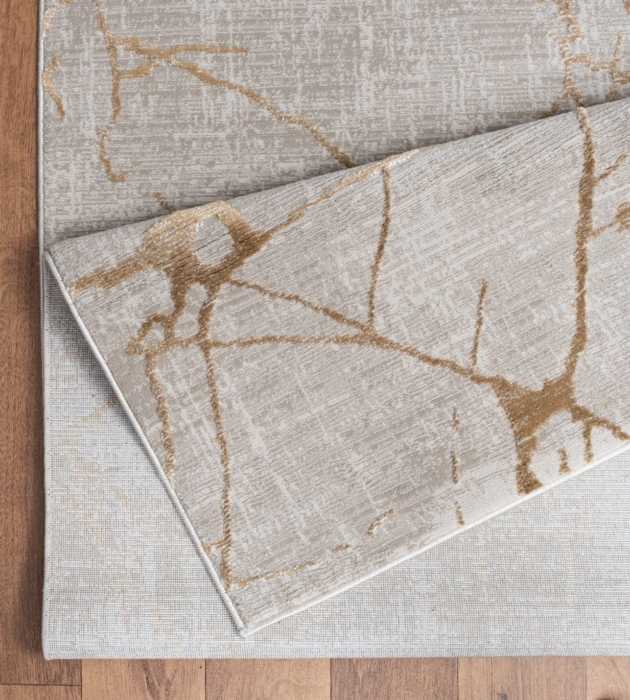 The Elegance - GC_CNC6004 Area Rug in beige features abstract gold and light gray lines, with one corner folded to show the underside. It is displayed on a wooden floor.