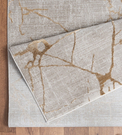 The Elegance - GC_CNC6004 Area Rug in beige features abstract gold and light gray lines, with one corner folded to show the underside. It is displayed on a wooden floor.