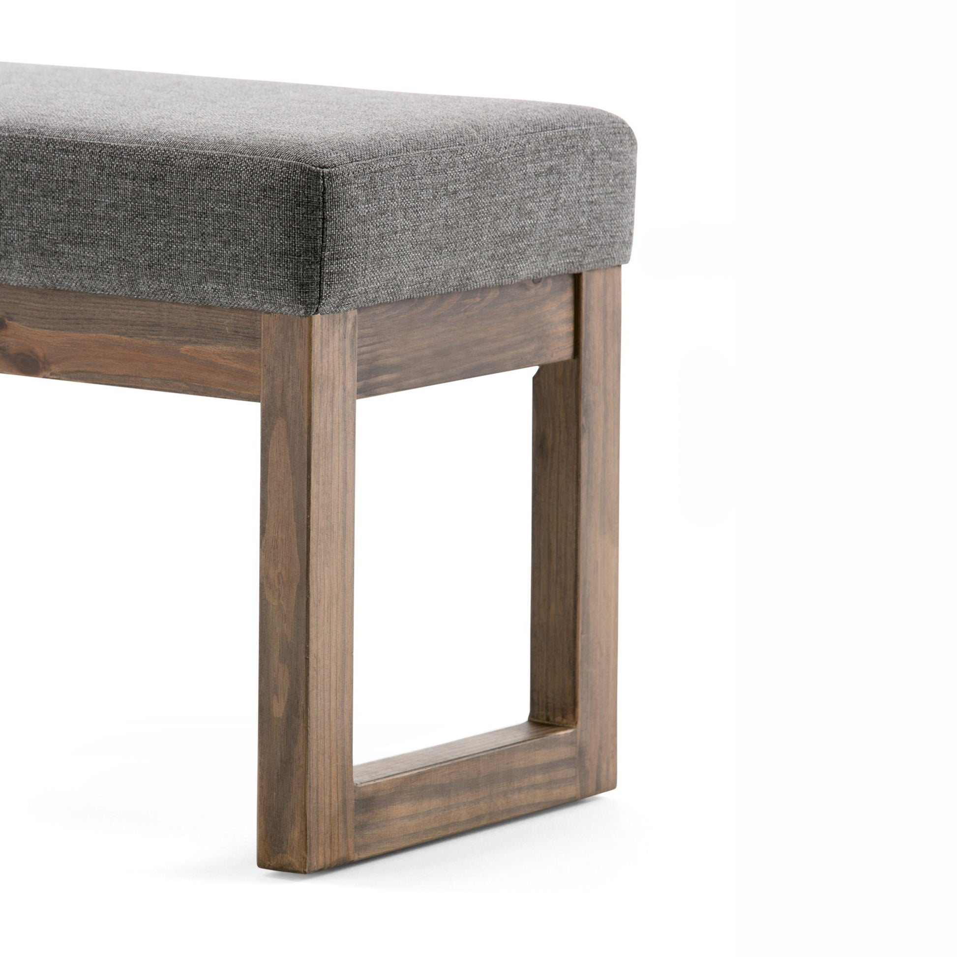 A close-up of the Milltown - Upholstered Ottoman Bench, featuring a rectangular grey seat and sturdy wooden legs in a geometric open-frame design, set against a white background.