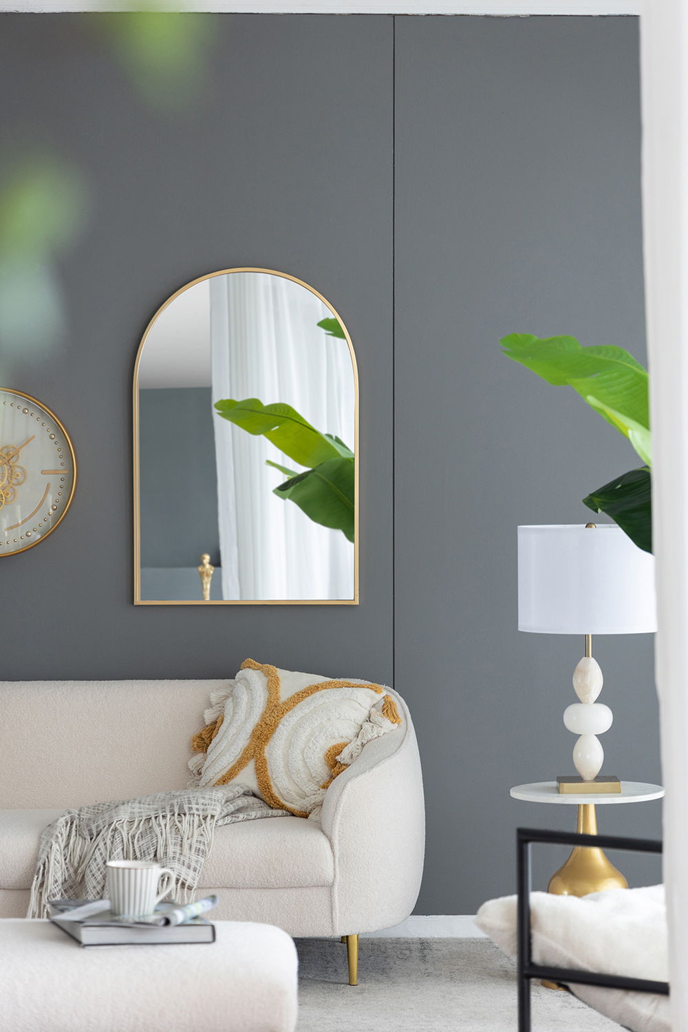 Modern living room with a gray accent wall, Arched Mirror With Metal Frame for living room, gold-framed clock, white couch with patterned pillows and throw, white table lamp, green plant, and natural light through the window.