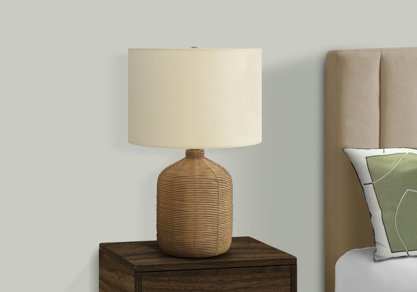 A modern rattan table lamp sits on a nightstand beside a bed with a tan upholstered headboard and a green and white decorative pillow.