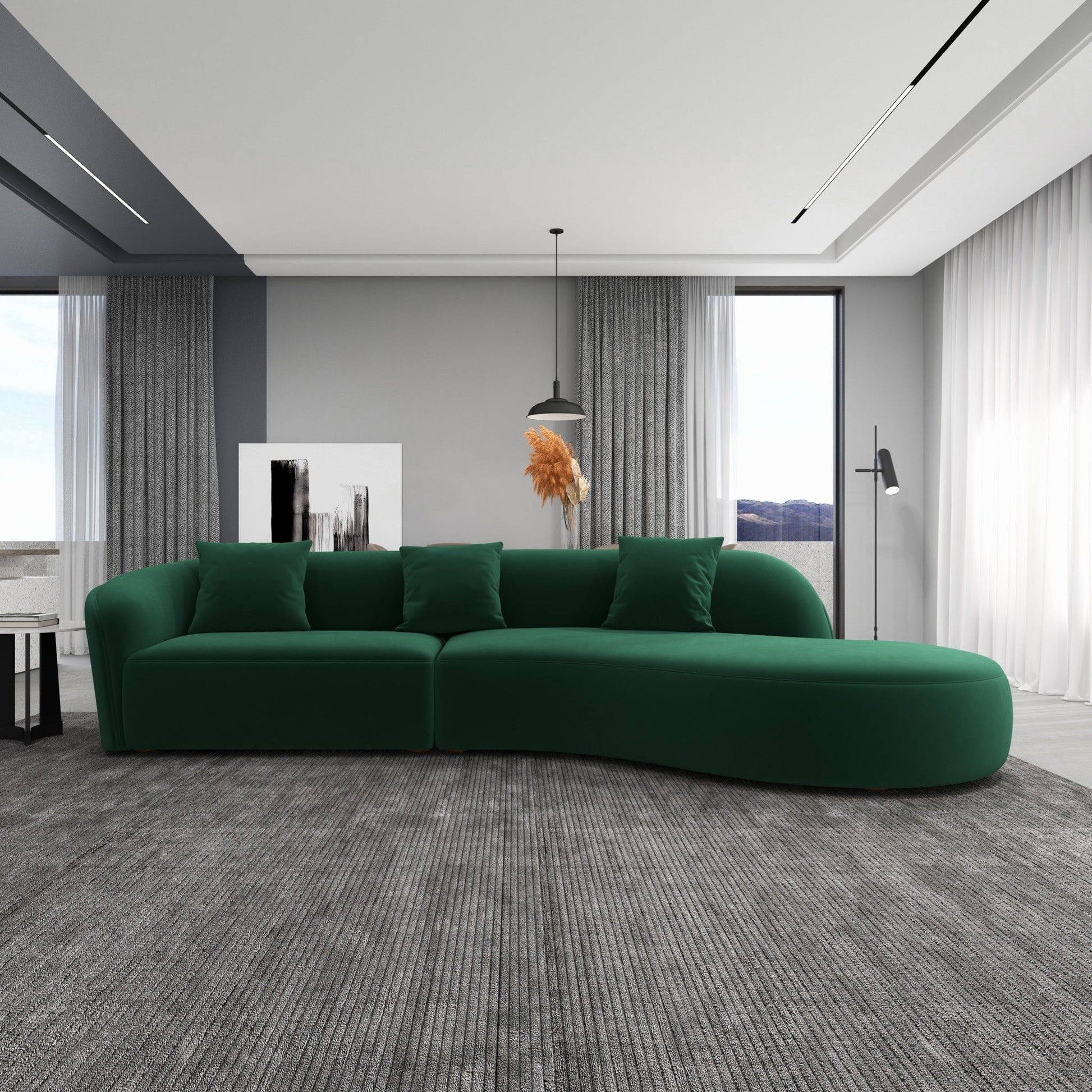 A modern living room featuring the Elijah Japandi Style Curvy Sectional Sofa in green with four cushions, a gray textured rug, sheer floor-to-ceiling curtains, gray walls, a pendant light, artwork, and a decorative plant.