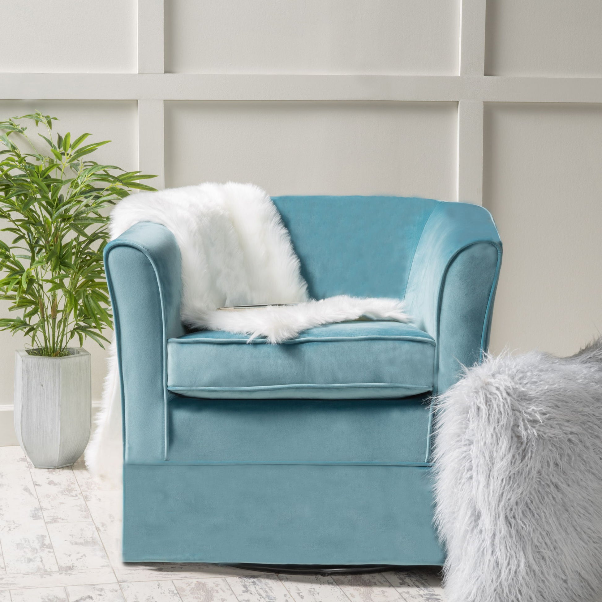 The Zeniqua Upholstered Velvet Swivel Chair with Loose Cover in light blue sits on a light wooden floor, draped with a white faux fur throw. A potted green plant is on the left and a fluffy gray rug peeks in from the right.