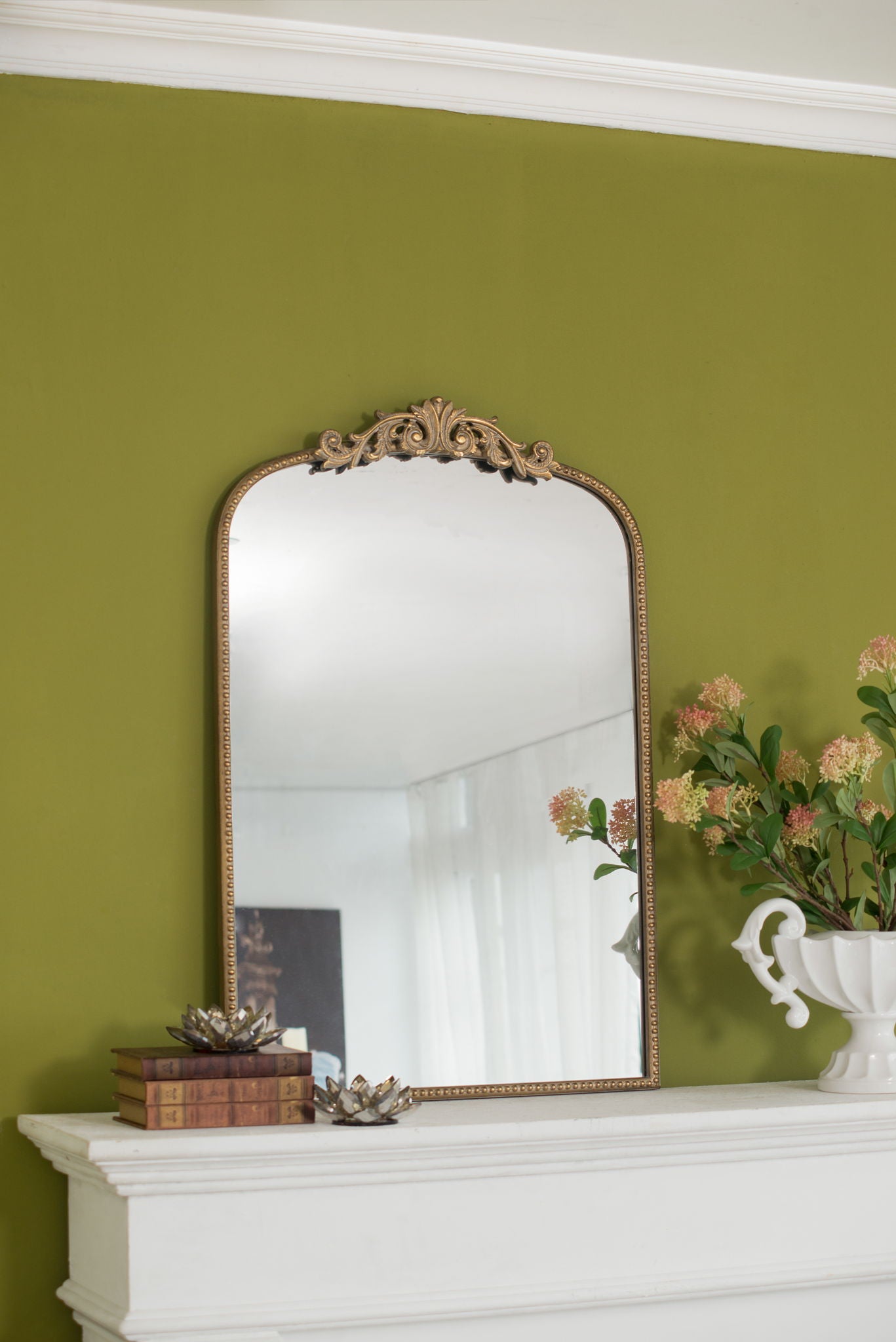 The Arch Mirror, Baroque Inspired Wall Decor, with an ornate gold frame sits on a white mantel against a green wall, accompanied by stacked books, a small decor piece, and a white vase filled with pink flowers.