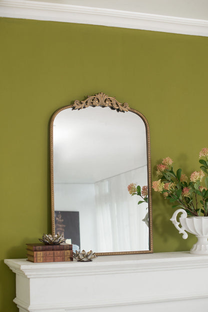 The Arch Mirror, Baroque Inspired Wall Decor, with an ornate gold frame sits on a white mantel against a green wall, accompanied by stacked books, a small decor piece, and a white vase filled with pink flowers.