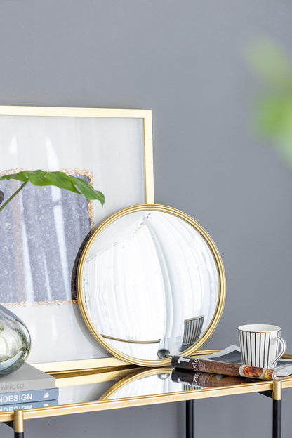 A Round Mirror with an iron frame leans against a framed picture on a glass table, surrounded by stacked books, a leafy plant in a vase, and a striped mug—all set before a gray wall. Ideal for living rooms, bedrooms, vanities, or entryways.