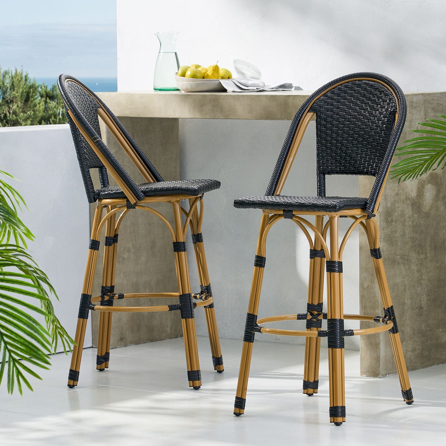 Two Outdoor Rattan and Aluminum French Barstools (Set of 2) with curved black woven seats and tan legs stand by a concrete outdoor counter topped with a pitcher, lemons, and plates. Green plants and blue sky are seen in the background.