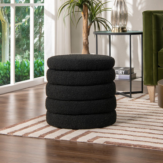 The Fuji Oversized Round Storage Ottoman, black and textured, sits on a striped rug in a modern living room with wood floors, a green velvet chair, side table, leafy plant, and sunlight streaming through the window.