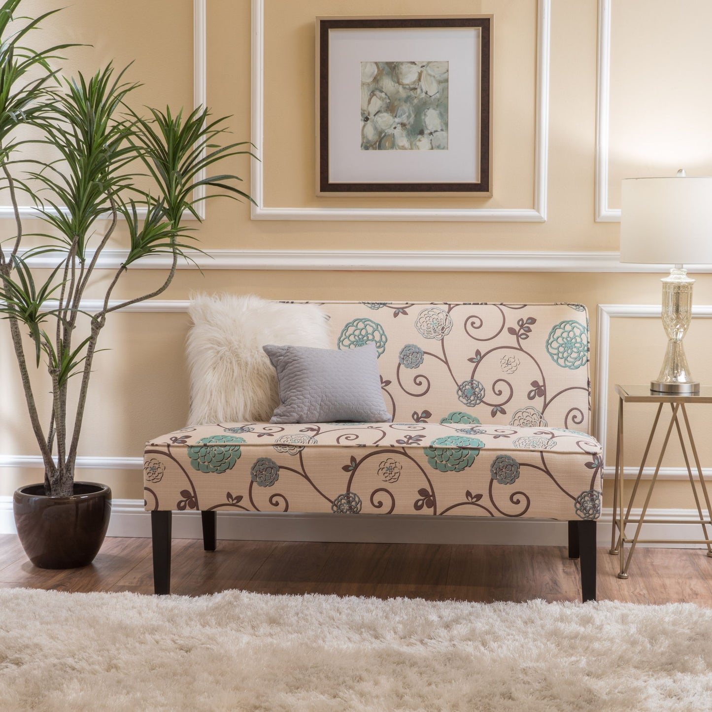 A cozy living room corner featuring a Contemporary Loveseat with upholstered fabric and birch wood legs, two pillows, a potted plant, framed art on a cream wall, a floor lamp, and a fluffy white rug on wooden flooring.