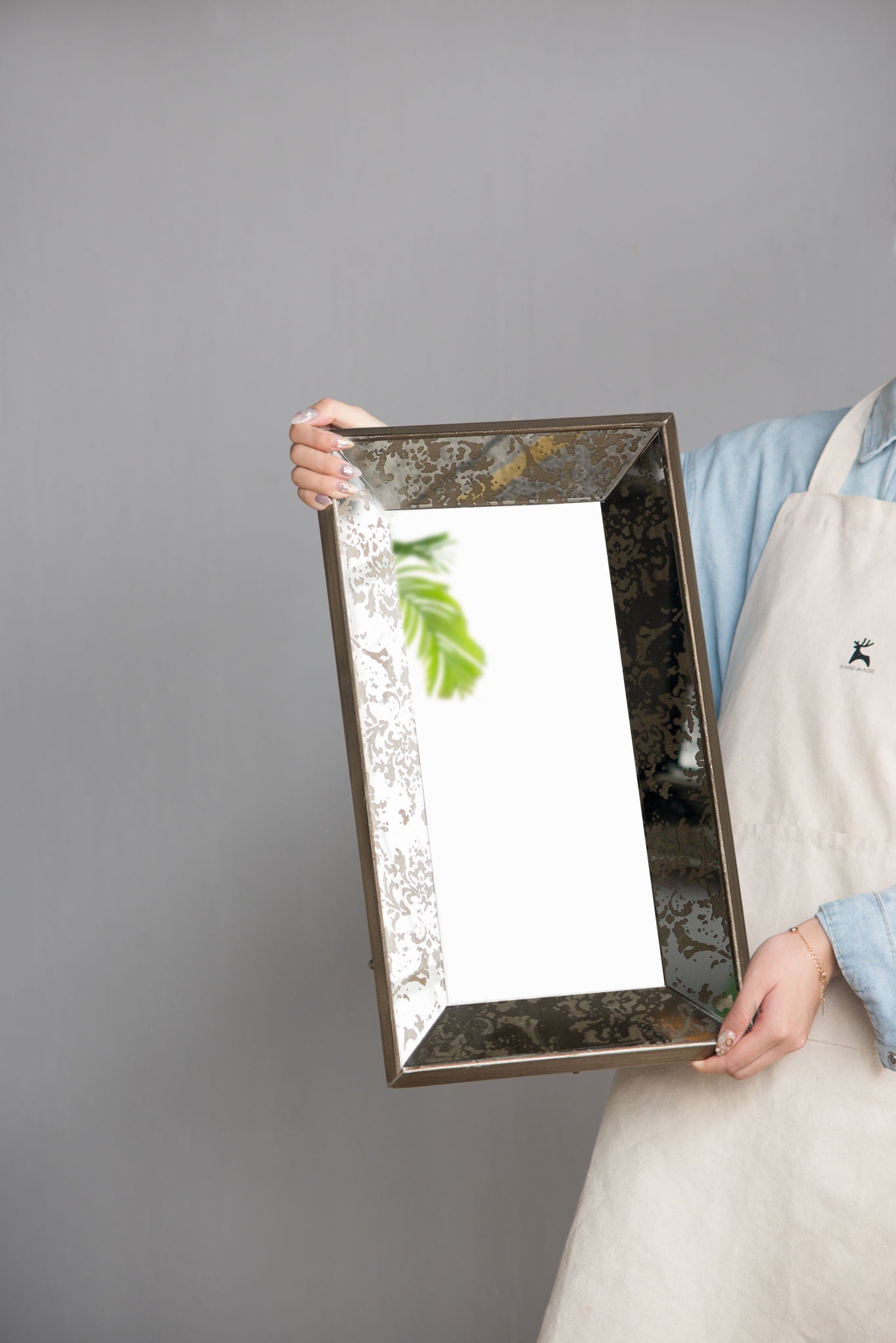 Against a plain gray background, a person wearing a light blue shirt and beige apron holds the Antique Rectangle Mirror With Floral Accents, Mirrored Display Tray, which reflects a leafy green plant.