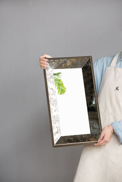 Against a plain gray background, a person wearing a light blue shirt and beige apron holds the Antique Rectangle Mirror With Floral Accents, Mirrored Display Tray, which reflects a leafy green plant.