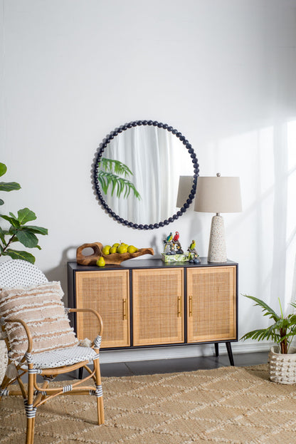 A cozy, modern room featuring a rattan chair with cushion, woven rug, black sideboard with wicker doors, plants, lemon accents, and sunlight through sheer curtains. On the wall hangs a Circle Mirror With Metal Beaded Frame in black.