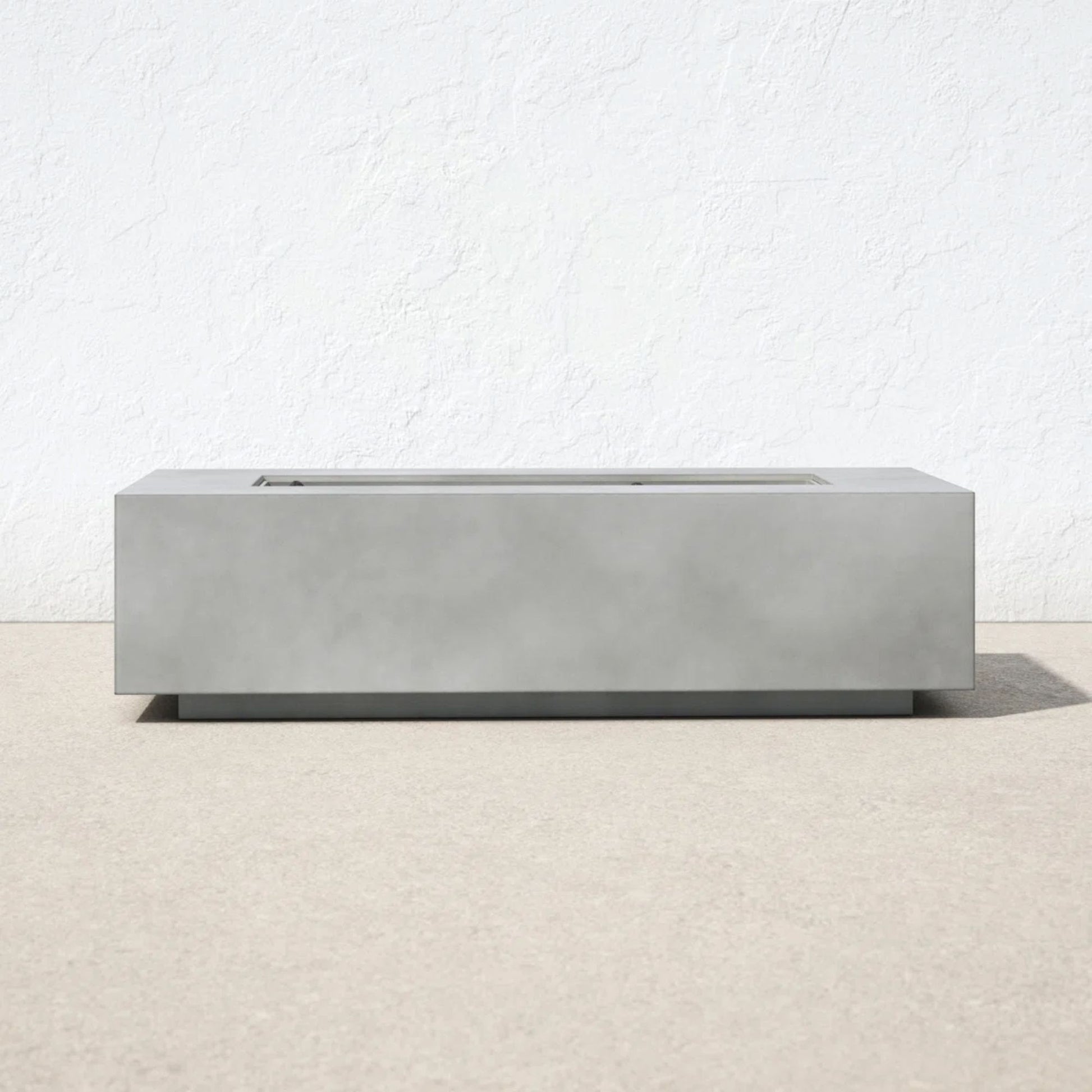 The Modern Concrete Design Propane Outdoor Fire Pit Table, featuring a minimalist rectangular shape, rests on a light beige surface with a plain white textured wall behind it. The design is sleek, with no visible flames or extra decorations.