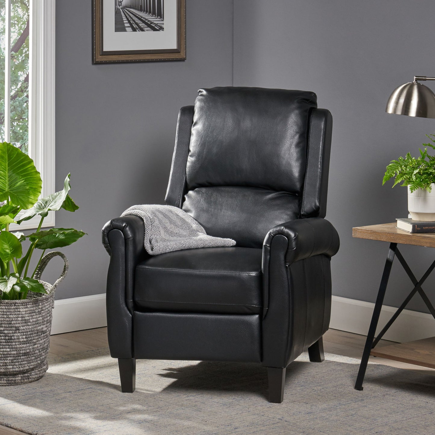 The Contemporary Push Back Recliner Chair in black leather, draped with a gray patterned blanket, sits on a light rug beside a green potted plant and a wooden side table with a lamp and another plant in a cozy gray room.