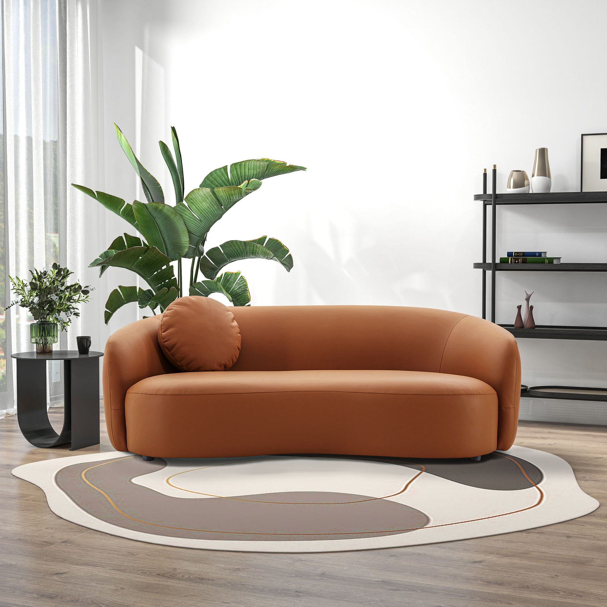 A modern living room featuring the Porter - Comfortable Sofa in brown, a round pillow, a large green plant, a black side table with a flower vase, a patterned rug, wooden floors, and a black shelving unit against a white wall.