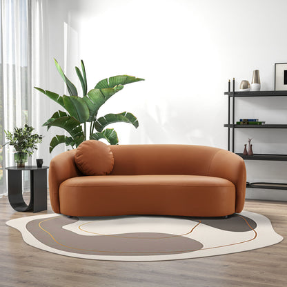A modern living room featuring the Porter - Comfortable Sofa in brown, a round pillow, a large green plant, a black side table with a flower vase, a patterned rug, wooden floors, and a black shelving unit against a white wall.